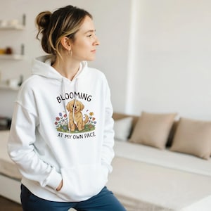 May include: White hoodie featuring a cartoon golden retriever puppy with flowers, and the text "BLOOMING AT MY OWN PACE". The hoodie has a front pocket and a drawstring hood. The image is on the front of the garment.