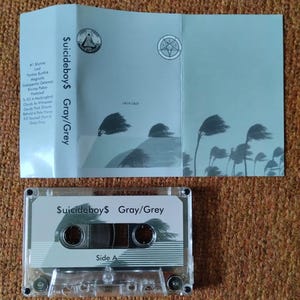 May include: A clear cassette tape and its accompanying light blue case are displayed. The cassette tape is labeled "$uicideboy$ Gray/Grey" with "Side A" visible. The case features the same title and track names, along with stylized illustrations.