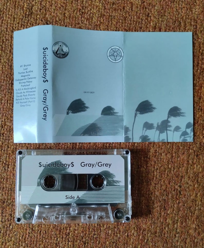May include: A clear cassette tape and its accompanying light blue case are displayed. The cassette tape is labeled "$uicideboy$ Gray/Grey" with "Side A" visible. The case features the same title and track names, along with stylized illustrations.