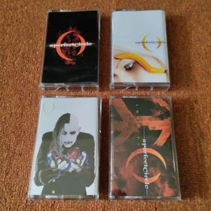 May include: Four cassette tapes with album art. The top left tape has a black background with a red and orange design and the text "a perfect circle". The top right tape has a white background with a woman's eye and a yellow object. The bottom left tape has a man with white and black hair holding a heart. The bottom right tape has a red and orange background with the text "a perfect circle".