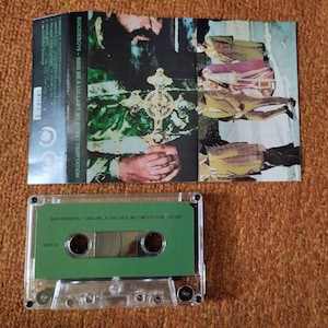 May include: A cassette tape with a green label and the text "SUICIDEBOYS - SING ME A LULLABY, MY SWEET TEMPTATION." The cassette case is clear. The album art features a religious figure holding a cross.