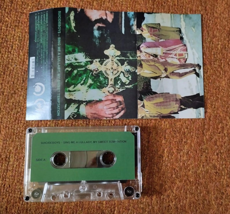May include: A cassette tape with a green label and the text "SUICIDEBOYS - SING ME A LULLABY, MY SWEET TEMPTATION." The cassette case is clear. The album art features a religious figure holding a cross.