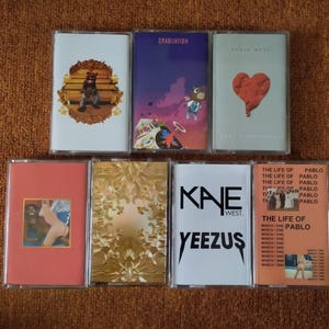 Könnte beinhalten: Eine Sammlung von acht Kassetten mit verschiedenen Albumcovern. Die Cover zeigen eine Vielzahl von Designs, darunter einen Bären, der auf einer Bank sitzt, einen Comic-Charakter, ein rotes Herz und ein goldenes Design. Der Text auf den Kassetten lautet "Graduation", "Kanye West", "KNE WEST", "YEEZUS" und "The Life of Pablo".