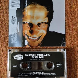 May include: A black cassette tape with a clear plastic case. The tape is labeled "Richard D. James Album: Aphex Twin" and "Warp MC43". The tape is a music recording from 1990.