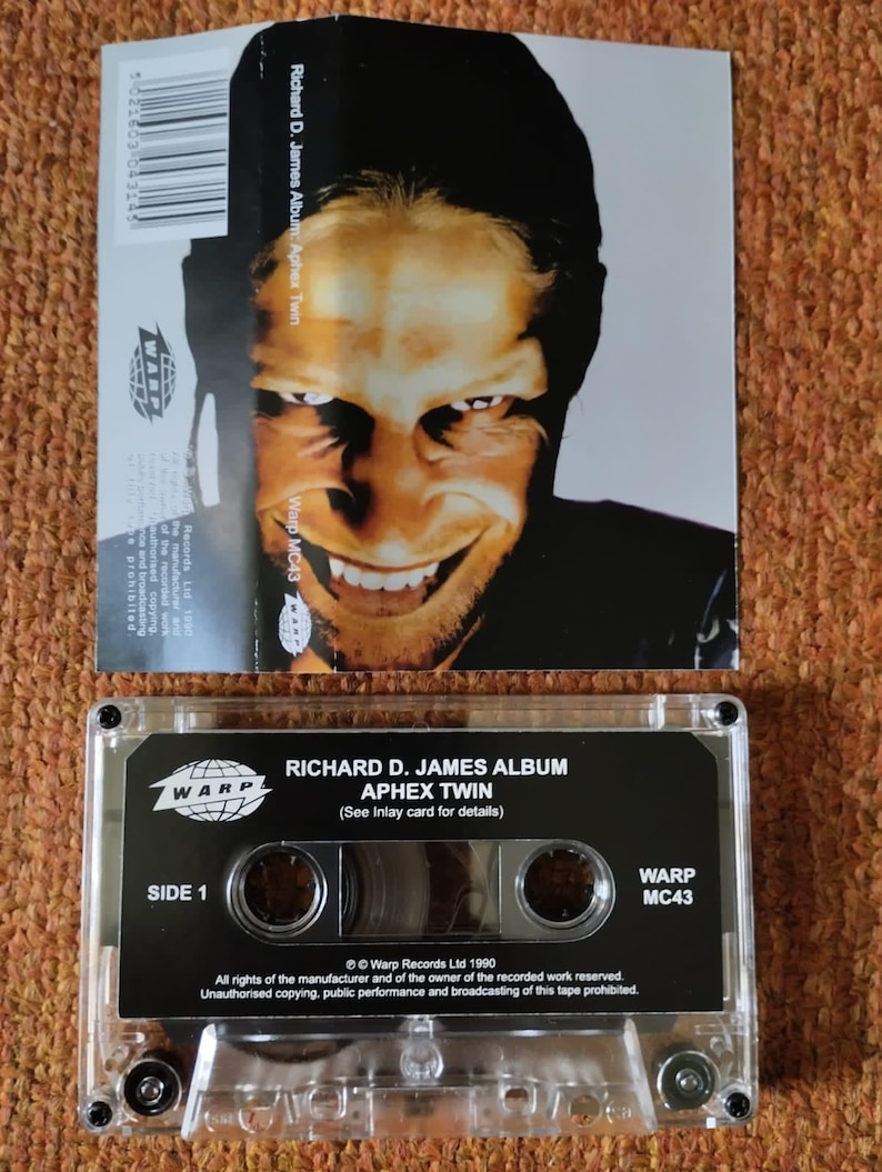 May include: A black cassette tape with a clear plastic case. The tape is labeled "Richard D. James Album: Aphex Twin" and "Warp MC43". The tape is a music recording from 1990.
