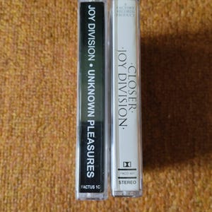 Puede incluir: Dos cintas de casete, una negra y otra blanca, con el nombre de la banda Joy Division y los t&iacute;tulos de los &aacute;lbumes impresos en ellas. La cinta negra est&aacute; etiquetada como "Joy Division &bull; Unknown Pleasures" y la cinta blanca est&aacute; etiquetada como "Closer &bull; Joy Division".