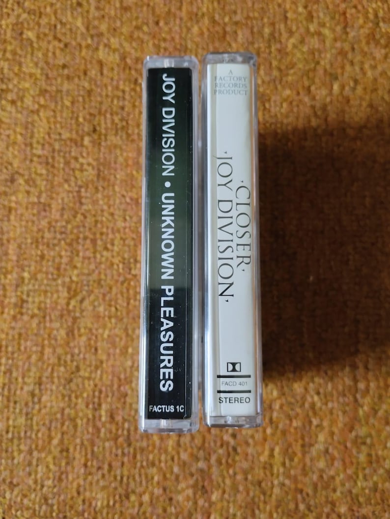 Puede incluir: Dos cintas de casete, una negra y otra blanca, con el nombre de la banda Joy Division y los t&iacute;tulos de los &aacute;lbumes impresos en ellas. La cinta negra est&aacute; etiquetada como "Joy Division &bull; Unknown Pleasures" y la cinta blanca est&aacute; etiquetada como "Closer &bull; Joy Division".
