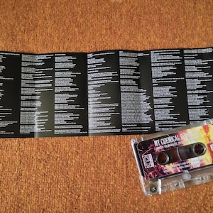 May include: A clear cassette tape with a colorful label featuring the text "MY CHEMICAL ROMANCE" and "THE BLACK PARADE". The cassette is next to a black and white lyric sheet on a brown surface.