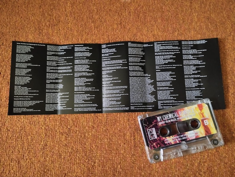 May include: A clear cassette tape with a colorful label featuring the text "MY CHEMICAL ROMANCE" and "THE BLACK PARADE". The cassette is next to a black and white lyric sheet on a brown surface.