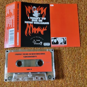 May include: An orange cassette tape and album art for "I Want to Die in New Orleans" by Suicideboys. The cassette tape has the album title and artist name printed on it. The album art features flames and the text "I Want to Die in New Orleans".