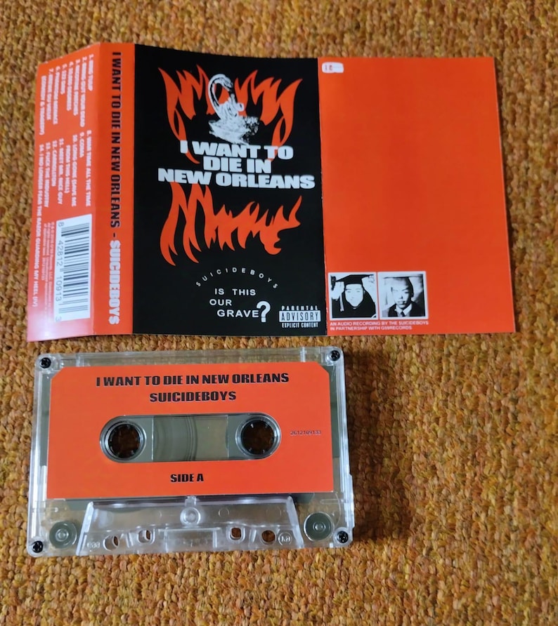 May include: An orange cassette tape and album art for "I Want to Die in New Orleans" by Suicideboys. The cassette tape has the album title and artist name printed on it. The album art features flames and the text "I Want to Die in New Orleans".