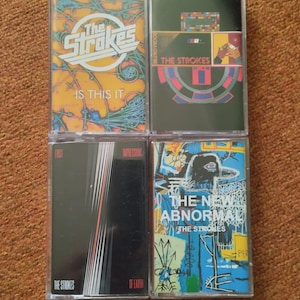 May include: Four cassette tapes by The Strokes. The tapes are in clear plastic cases with colourful artwork. The titles of the tapes are "Is This It", "Room On Fire", "First Impressions of Earth", and "The New Abnormal".