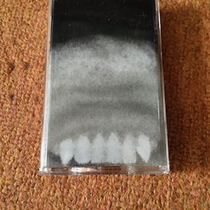 May include: A clear cassette tape case featuring an abstract black and white design. The design fades from black at the top to white at the bottom, resembling clouds. The cassette tape is placed on a brown, textured surface.