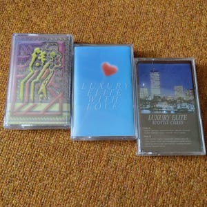 May include: Three cassette tapes with different designs. The first tape has a colorful abstract design. The second tape has a blue background with a white heart and the text "Luxury Elite With Love". The third tape has a cityscape at night with the text "Luxury Elite World Class".