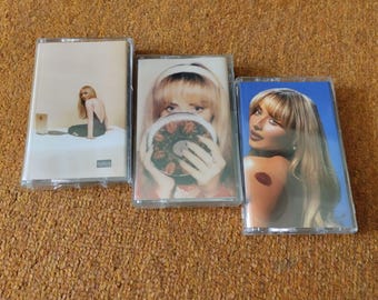 May include: Three cassette tapes with album art. The first shows a woman sitting on a bed. The second features a woman holding a mirror. The third has a woman with a red lip tattoo on her shoulder, with a blue background.
