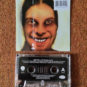 May include: A black cassette tape with a clear plastic case. The tape is labeled "aphex twin...I care because you do". The label also includes the track list and credits for the album. The tape is a physical copy of the album "I care because you do" by the electronic music artist Aphex Twin.
