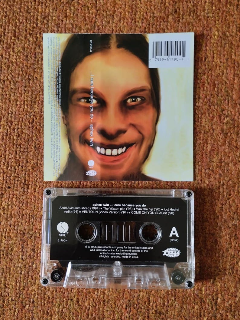 May include: A black cassette tape with a clear plastic case. The tape is labeled "aphex twin...I care because you do". The label also includes the track list and credits for the album. The tape is a physical copy of the album "I care because you do" by the electronic music artist Aphex Twin.