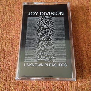 Puede incluir: Una cinta de casete negra con un gr&aacute;fico blanco de un patr&oacute;n de onda. El texto "JOY DIVISION" y "UNKNOWN PLEASURES" est&aacute; impreso en blanco en la cinta.