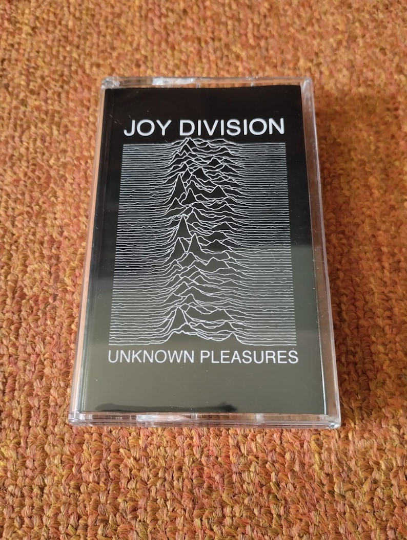 Puede incluir: Una cinta de casete negra con un gr&aacute;fico blanco de un patr&oacute;n de onda. El texto "JOY DIVISION" y "UNKNOWN PLEASURES" est&aacute; impreso en blanco en la cinta.