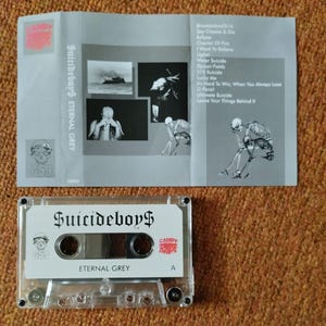 May include: A clear cassette tape labeled "Suicideboy$" and "Eternal Grey" with a skull graphic. The cassette case is next to a gray folded insert with song titles and images. The insert has the text "Suicideboy$ Eternal Grey".