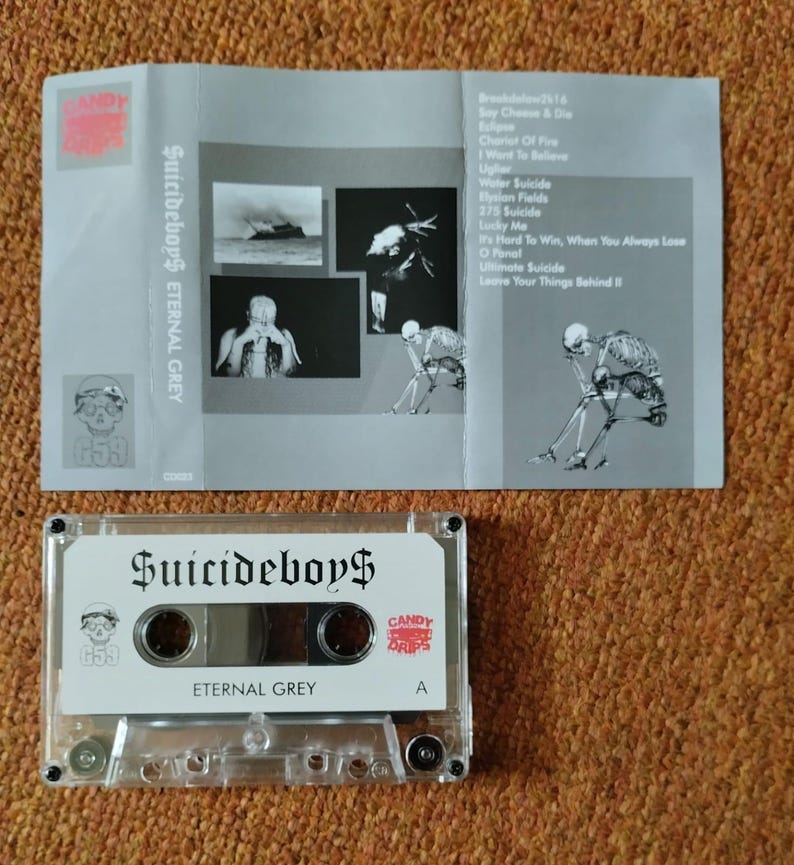 May include: A clear cassette tape labeled "Suicideboy$" and "Eternal Grey" with a skull graphic. The cassette case is next to a gray folded insert with song titles and images. The insert has the text "Suicideboy$ Eternal Grey".