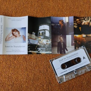 以下が含まれることがあります： 白いラベルに「Lost In Translation」と書かれた透明なカセットテープ。その隣には、映画のスチール写真とタイトルが掲載された折り畳まれた小冊子があります。アイテムはオレンジ色のテクスチャのある表面にあります。