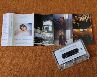 May include: A clear cassette tape labeled "Lost In Translation" with a white label and black text. The cassette is next to a folded booklet with movie stills and the film's title. The background is a textured orange surface.