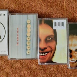 May include: Four cassette tapes by Aphex Twin. The first tape is white with a black logo. The second tape has a blue and white background with the text "The Aphex Twin" and "CLASSICS" in white. The third tape has a photo of a man with a large smile and the text "Richard D. James Album." The fourth tape has a photo of a man with a dark background and the text "Selected Ambient Works 85-92."