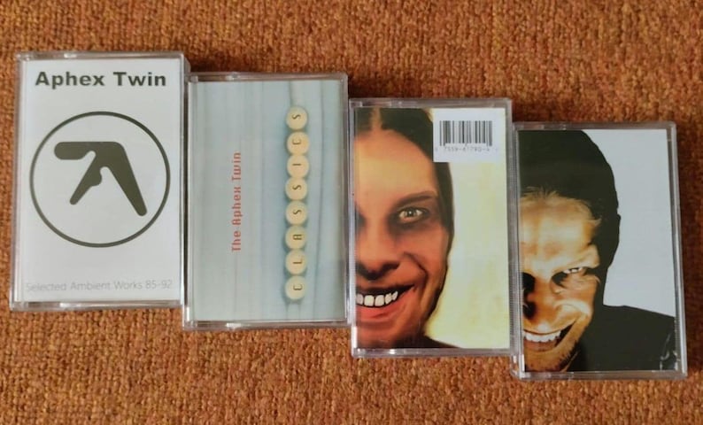 May include: Four cassette tapes by Aphex Twin. The first tape is white with a black logo. The second tape has a blue and white background with the text "The Aphex Twin" and "CLASSICS" in white. The third tape has a photo of a man with a large smile and the text "Richard D. James Album." The fourth tape has a photo of a man with a dark background and the text "Selected Ambient Works 85-92."