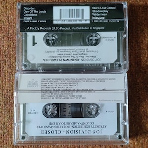 Puede incluir: Una cinta de casete negra con una etiqueta blanca. La etiqueta tiene el texto "Joy Division Closer" y "Atrocity Exhibition-Isolation-Passover-Colony A Means To An End" impreso en ella. La cinta es un producto de Factory Records.