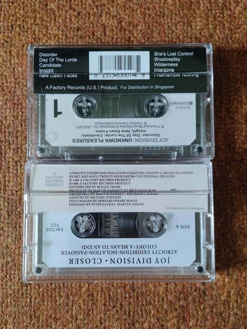 Puede incluir: Una cinta de casete negra con una etiqueta blanca. La etiqueta tiene el texto "Joy Division Closer" y "Atrocity Exhibition-Isolation-Passover-Colony A Means To An End" impreso en ella. La cinta es un producto de Factory Records.