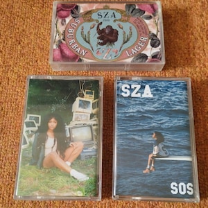 May include: Three cassette tapes featuring the artist SZA. The first tape is pink and white with a floral design and the text "SZA 1999 Suburban Lager". The second tape has a photo of SZA sitting in a grassy area with a pile of old televisions behind her. The third tape has a photo of SZA sitting on a pier over a body of water with the text "SOS".