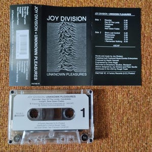 Puede incluir: Una cinta de casete en blanco y negro con el t&iacute;tulo "Joy Division - Unknown Pleasures" impreso en la etiqueta. La cinta de casete est&aacute; en una caja de pl&aacute;stico transparente con una etiqueta blanca que tiene el n&uacute;mero "1" impreso en ella.