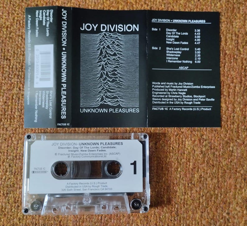 Puede incluir: Una cinta de casete en blanco y negro con el t&iacute;tulo "Joy Division - Unknown Pleasures" impreso en la etiqueta. La cinta de casete est&aacute; en una caja de pl&aacute;stico transparente con una etiqueta blanca que tiene el n&uacute;mero "1" impreso en ella.