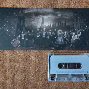 May include: A black and white photograph of a band on stage, with a cassette tape of the album "The Black Parade" by My Chemical Romance. The photo shows the band members in black clothing, with a drummer in the background. The cassette tape is in a clear plastic case with a black label.