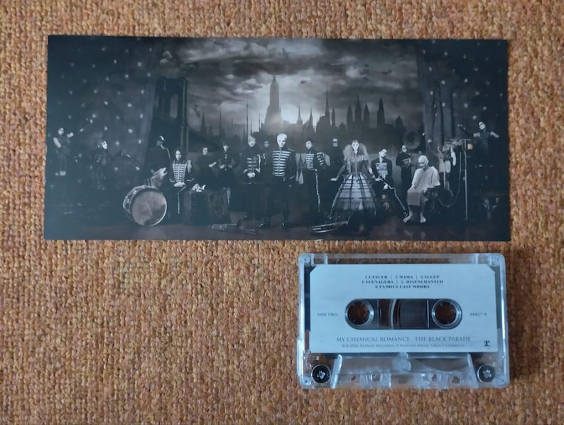 May include: A black and white photograph of a band on stage, with a cassette tape of the album "The Black Parade" by My Chemical Romance. The photo shows the band members in black clothing, with a drummer in the background. The cassette tape is in a clear plastic case with a black label.