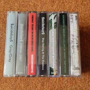 May include: A collection of seven cassette tapes with various album titles and the band name "$uicideboy$" printed on the spines. The tapes have different colored cases and labels, including gray, red, and black.