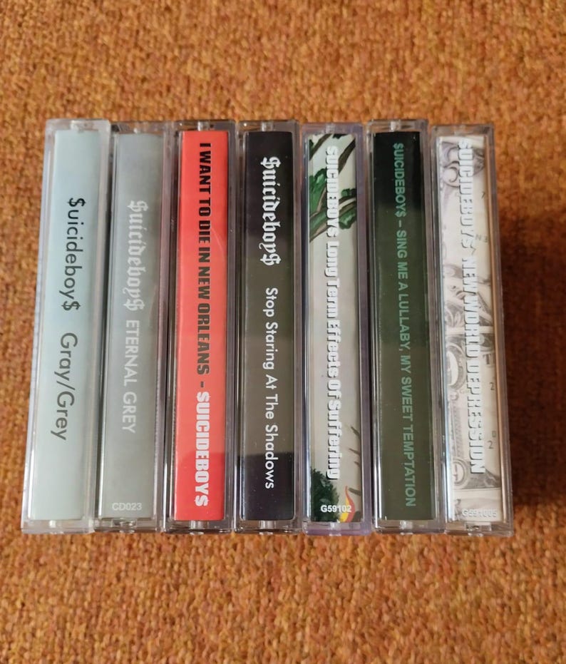 May include: A collection of seven cassette tapes with various album titles and the band name "$uicideboy$" printed on the spines. The tapes have different colored cases and labels, including gray, red, and black.