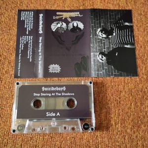May include: A clear cassette tape with the text "Suicideboy$ Stop Staring At The Shadows" and "Side A". The cassette case is open, revealing the album art with the band's name and album title. The album art is dark with images of the band.