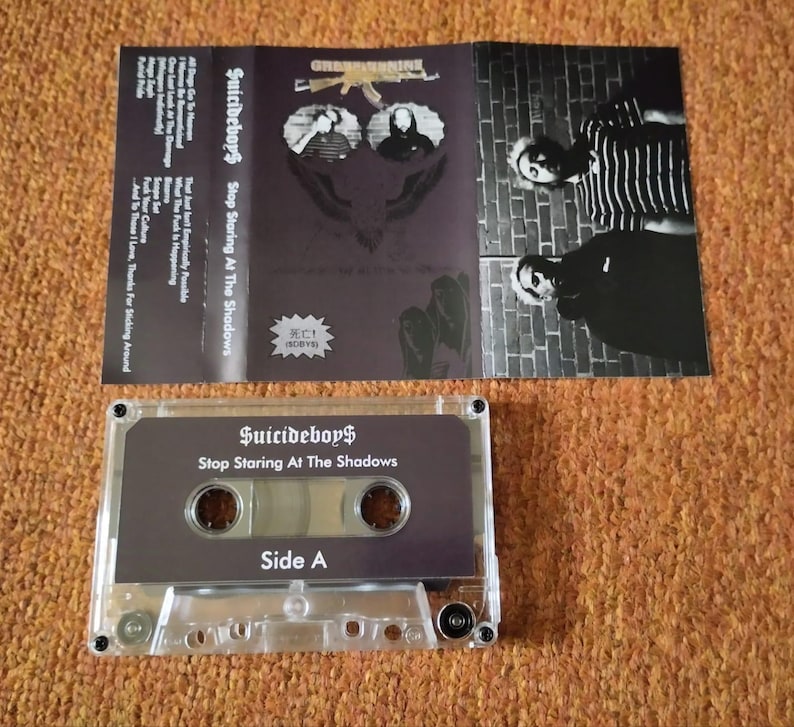 May include: A clear cassette tape with the text "Suicideboy$ Stop Staring At The Shadows" and "Side A". The cassette case is open, revealing the album art with the band's name and album title. The album art is dark with images of the band.