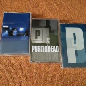 Può includere: Tre musicassette con copertine di album. La prima è blu scuro con una foto e la parola "DUMMY". La seconda è in bianco e nero con "PORTISHEAD". La terza è blu con una "P" bianca.