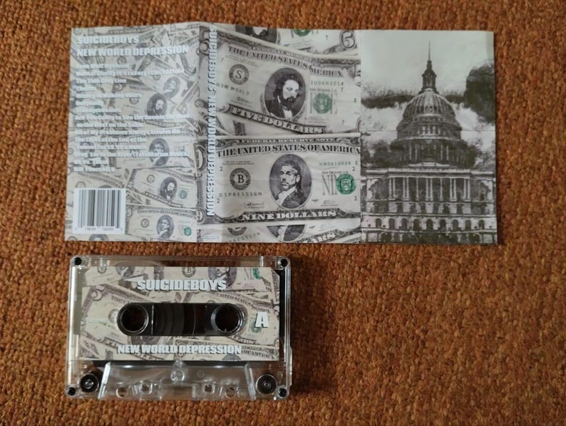 May include: A cassette tape and its cover featuring a design of US dollar bills. The cassette tape has the text "SUICIDEBOYS" and "NEW WORLD DEPRESSION". The cover also displays the text "SUICIDEBOYS" and a grayscale image of a building.