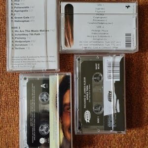 May include: Four clear cassette tapes with labels. The tapes are labeled with the names of the artists and albums. The tapes are on a brown surface.