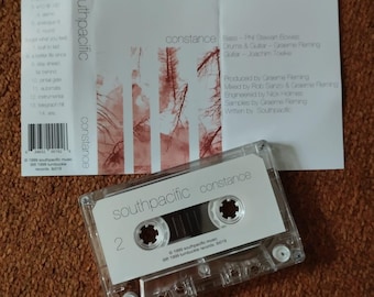May include: A clear cassette tape labelled "southpacific constance" with a white label and the number 2. The cassette case is next to a folded paper with the same title and artist information. The background is a brown, textured surface.