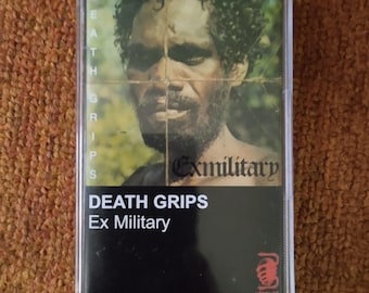 May include: A black cassette tape with a photo of a man on the front. The tape is titled "Death Grips Ex Military" and features a red scorpion logo.