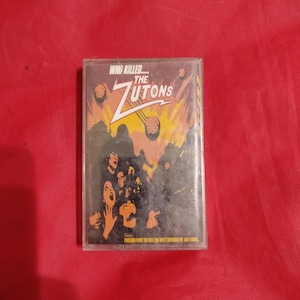 Op de afbeelding: Een vintage cassettebandje voor het album "The Zutons", met de tekst "Who Killed... The Zutons". De hoes toont een gestileerde illustratie in oranje, geel en zwart, met een groep mensen met geschokte uitdrukkingen.