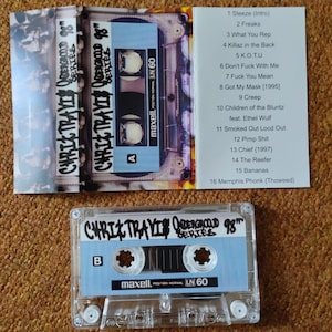May include: A clear cassette tape labeled "CHRIZ TRAVIS ODERGROUND SERIES 98" with a light blue label and the text "maxell LN 60." The cassette case is open, revealing the tracklist. The cassette tape is on a brown surface.