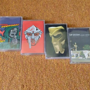 Puede incluir: Cuatro cintas de casete con ilustraciones coloridas. Las cintas presentan imágenes de una figura enmascarada y texto, incluyendo "DOOMSDAY" y "MF DOOM". Las carcasas de los casetes son de plástico transparente, sobre un fondo marrón texturizado.