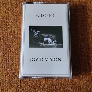 Puede incluir: Una cinta de casete blanca con una foto en blanco y negro de un hombre y una mujer en la parte delantera. El texto "CLOSER" y "JOY DIVISION" est&aacute; impreso en la cinta.