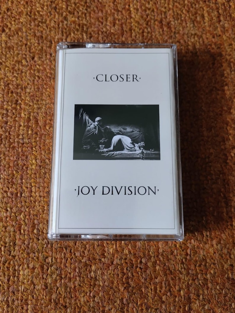 Puede incluir: Una cinta de casete blanca con una foto en blanco y negro de un hombre y una mujer en la parte delantera. El texto "CLOSER" y "JOY DIVISION" est&aacute; impreso en la cinta.